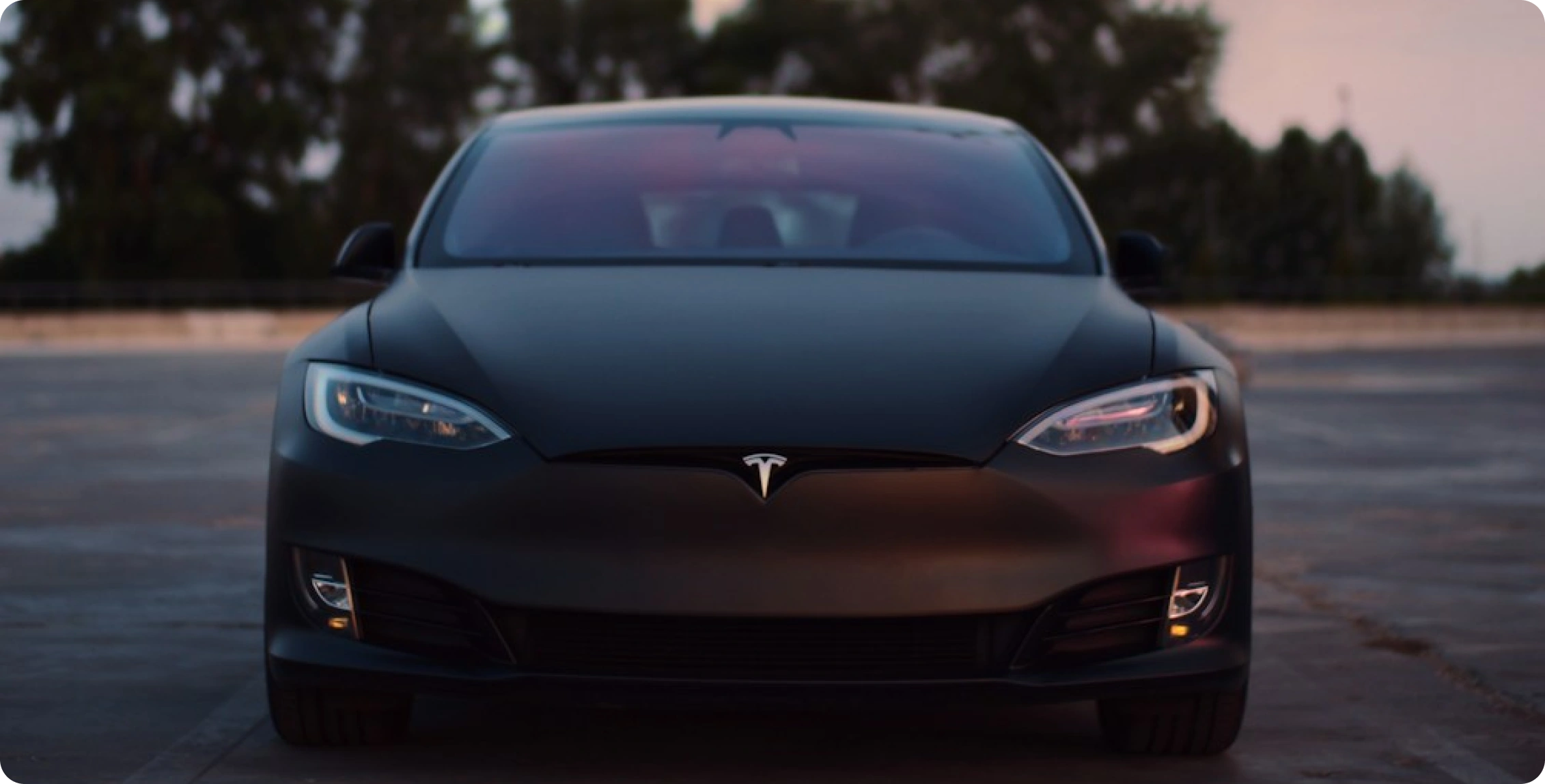 Čelní pohled na černý elektromobil Tesla zaparkovaný na prázdném parkovišti za soumraku, v pozadí jsou stromy.