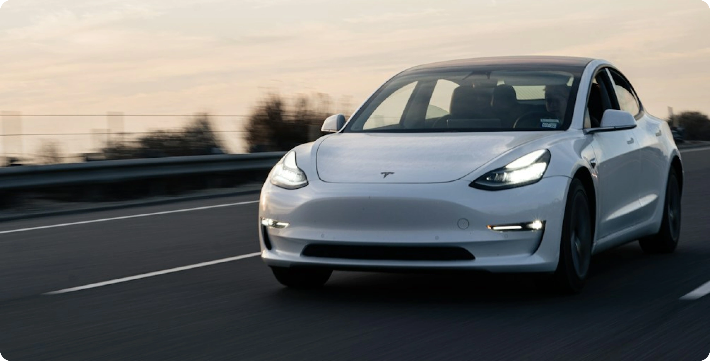 Bílý elektromobil Tesla jede po dálnici za soumraku – záběr zepředu se světly a lehkým rozmazáním pohybu