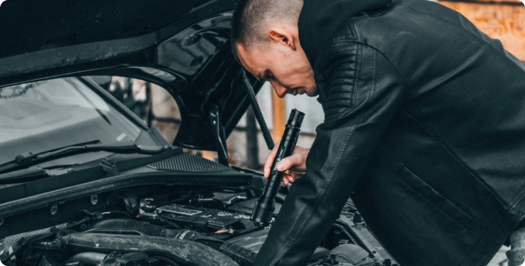Mechanik kontroluje motor auta pod otevřenou kapotou a svítí si baterkou