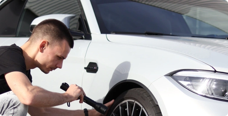 Mechanik kontroluje kolo bílého auta a svítí si baterkou u předního blatníku